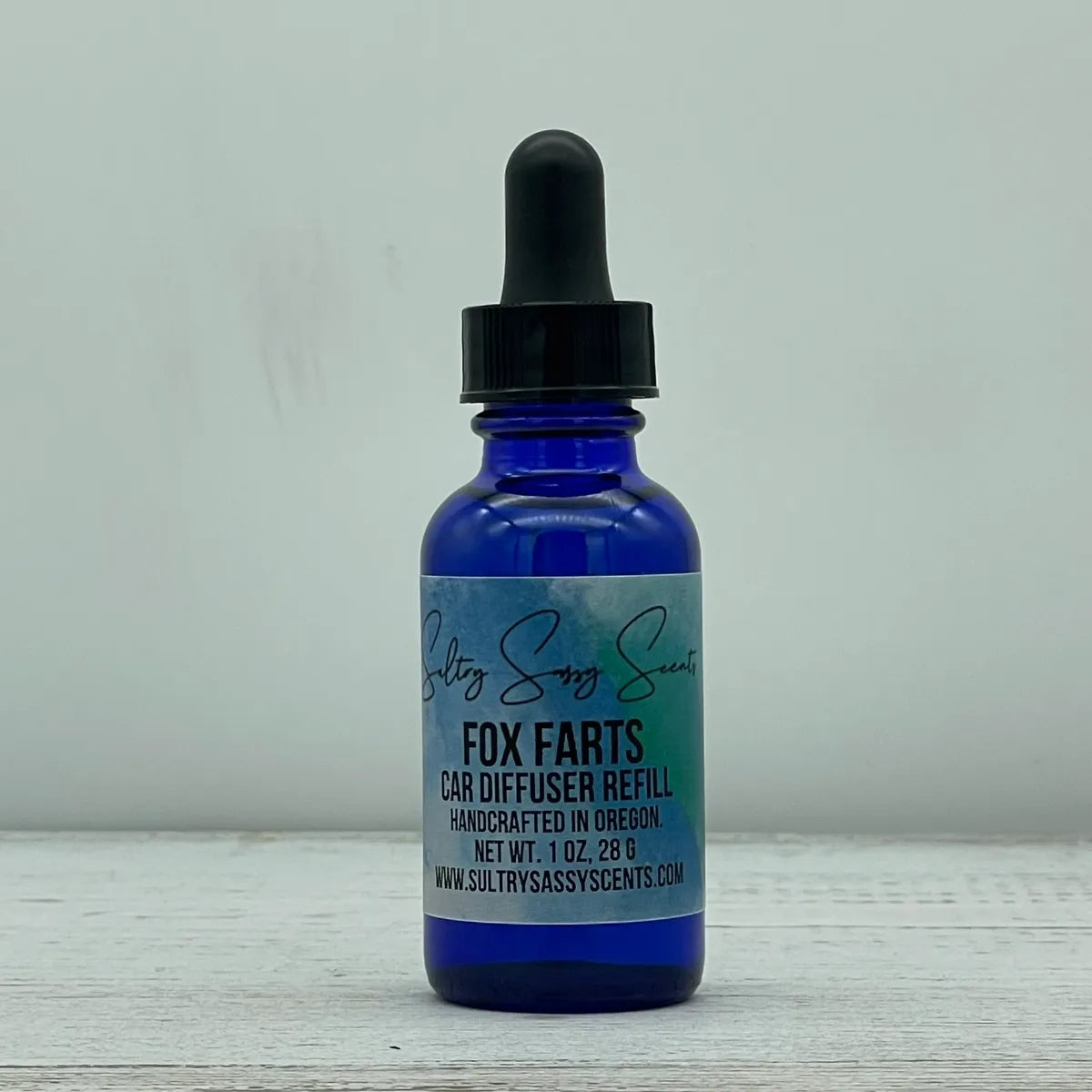 Front label view of a cobalt blue glass dropper bottle containing Sultry Sassy Scents "Fox Farts" Car Diffuser Refill, highlighting the text showing it is handcrafted in Oregon.