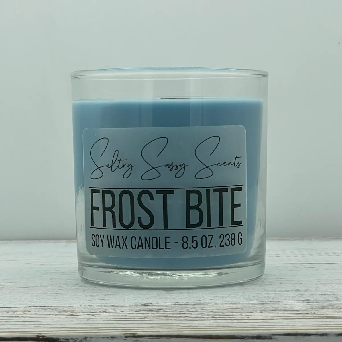 An 8.5-ounce clear glass jar of Sultry Sassy Scents "Frost Bite" soy wax candle featuring a smooth cornflower blue wax color, displayed on a rustic light wood table.