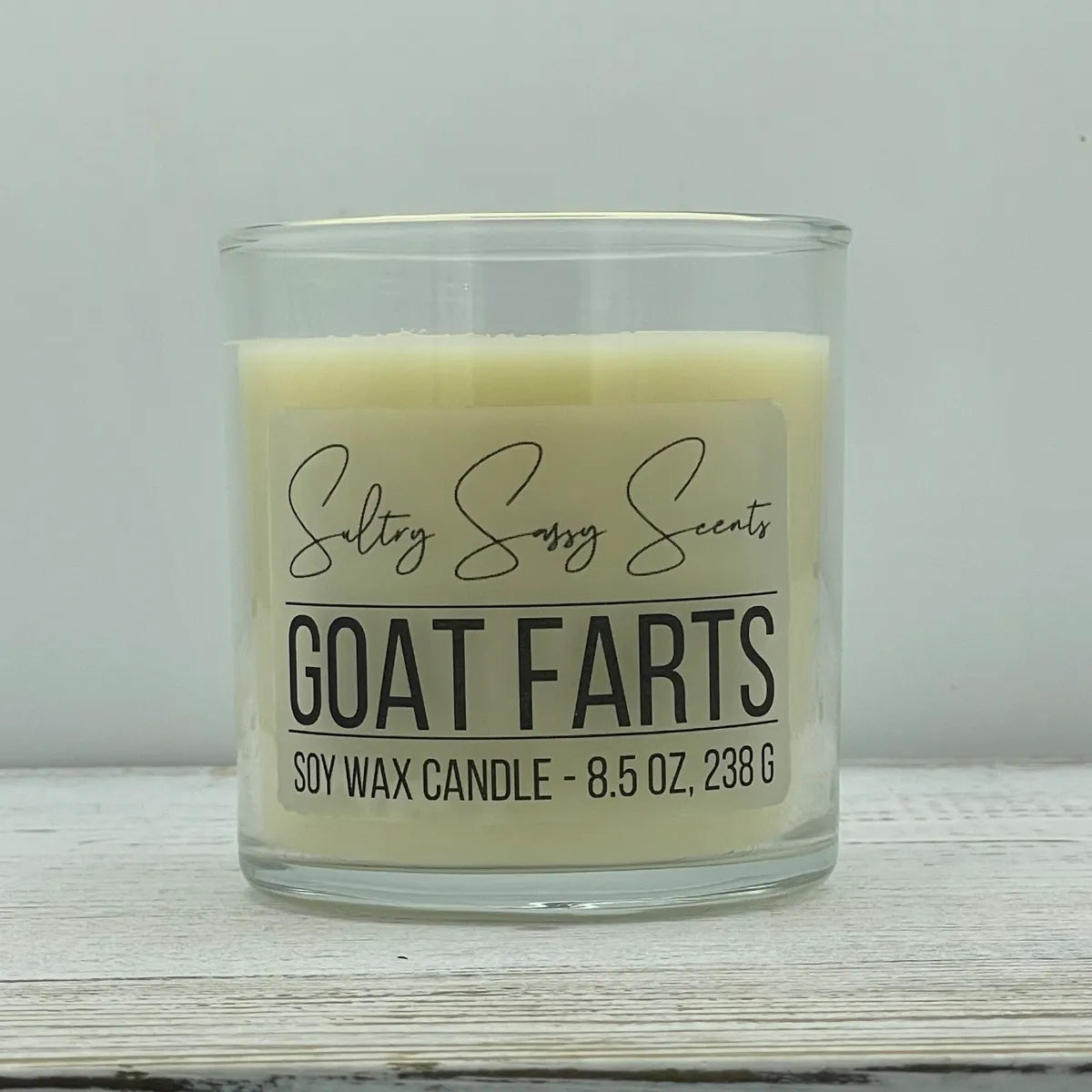 An 8.5-ounce clear glass jar of Sultry Sassy Scents "Goat Farts" soy wax candle showcasing a smooth creamy off-white wax color and a flat wooden wick.