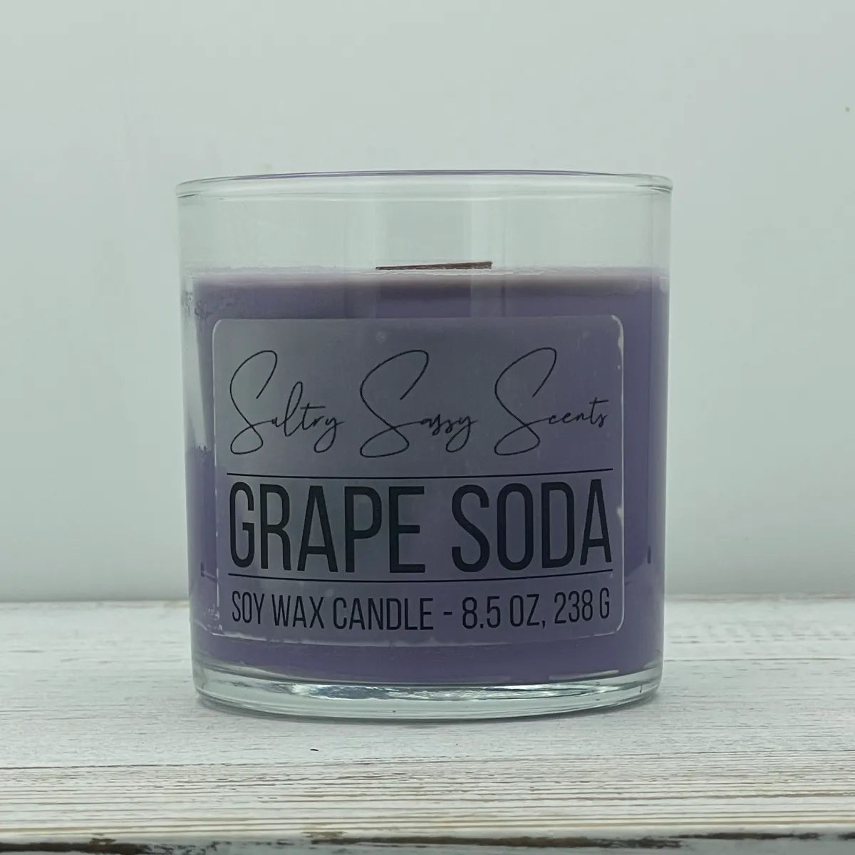 An 8.5-ounce clear glass jar of Sultry Sassy Scents "Grape Soda" soy wax candle featuring a dusty purple wax color and a flat wooden wick, resting on a white-washed wood table.