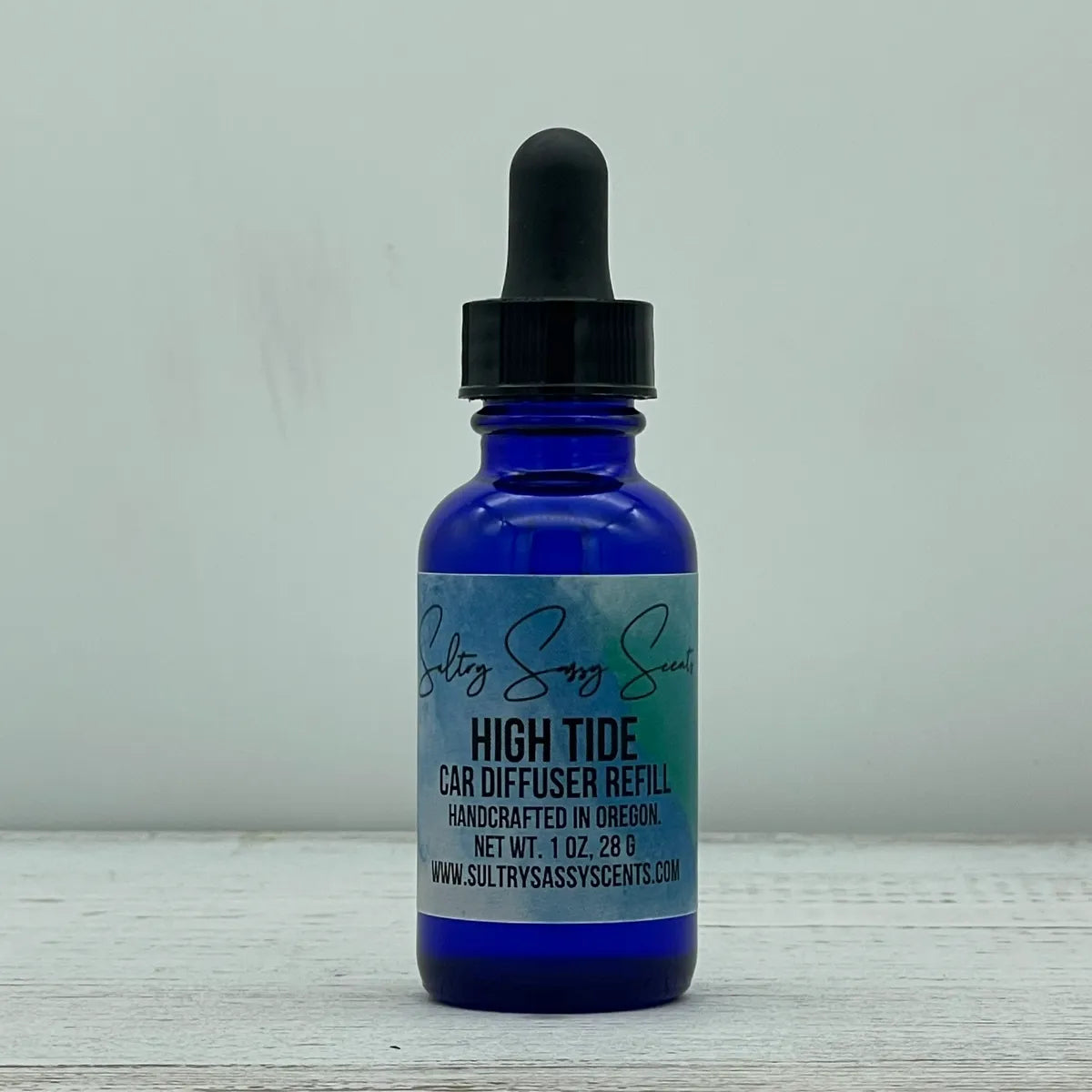 Front label view of a cobalt blue glass dropper bottle containing Sultry Sassy Scents "High Tide" Car Diffuser Refill, positioned on a rustic light wood table.