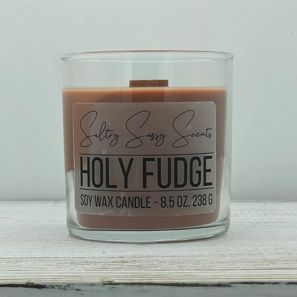 An 8.5-ounce clear glass jar of Sultry Sassy Scents "Holy Fudge" soy wax candle showcasing a rich milk chocolate brown wax color and a flat wooden wick.