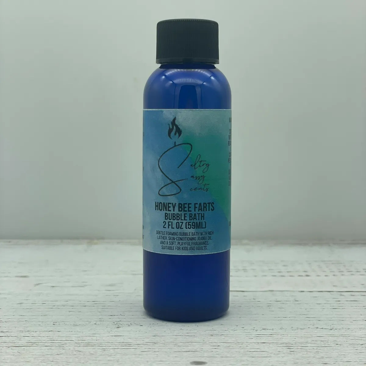 Front label view of a small 2-ounce travel size blue bottle with a black screw cap containing Sultry Sassy Scents "Honey Bee Farts" Bubble Bath, centered on a light wood backdrop.