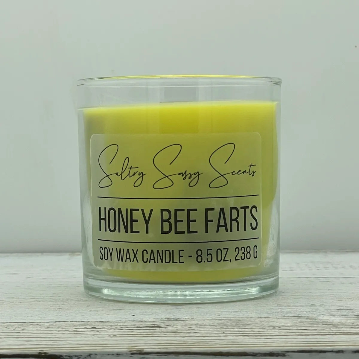 An 8.5-ounce clear glass jar of Sultry Sassy Scents "Honey Bee Farts" soy wax candle displaying a vibrant canary yellow wax color, centered on a white wood backdrop.