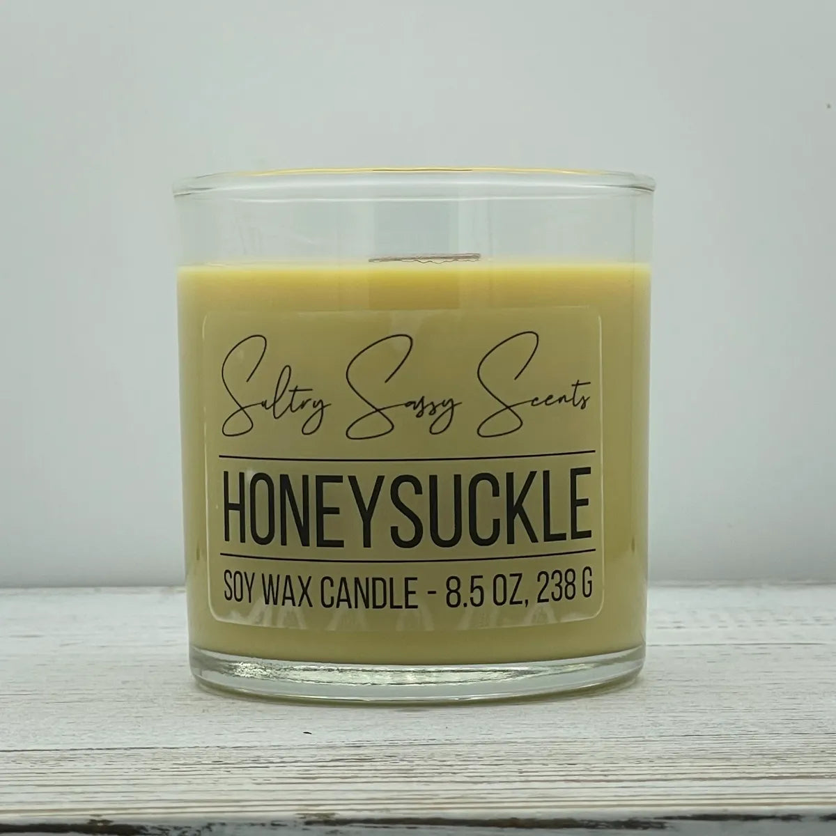 An 8.5-ounce clear glass jar of Sultry Sassy Scents "Honeysuckle" soy wax candle featuring a smooth, pale pastel yellow wax color and a flat wooden wick, centered on a white wood surface.