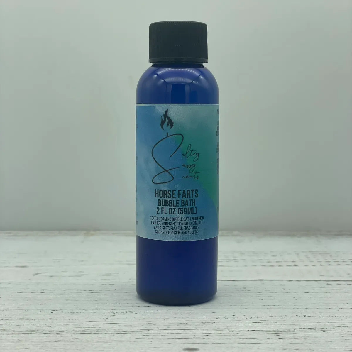 Front label view of a small 2-ounce travel size blue bottle with a black screw cap containing Sultry Sassy Scents "Horse Farts" Bubble Bath, resting on a white wood surface.