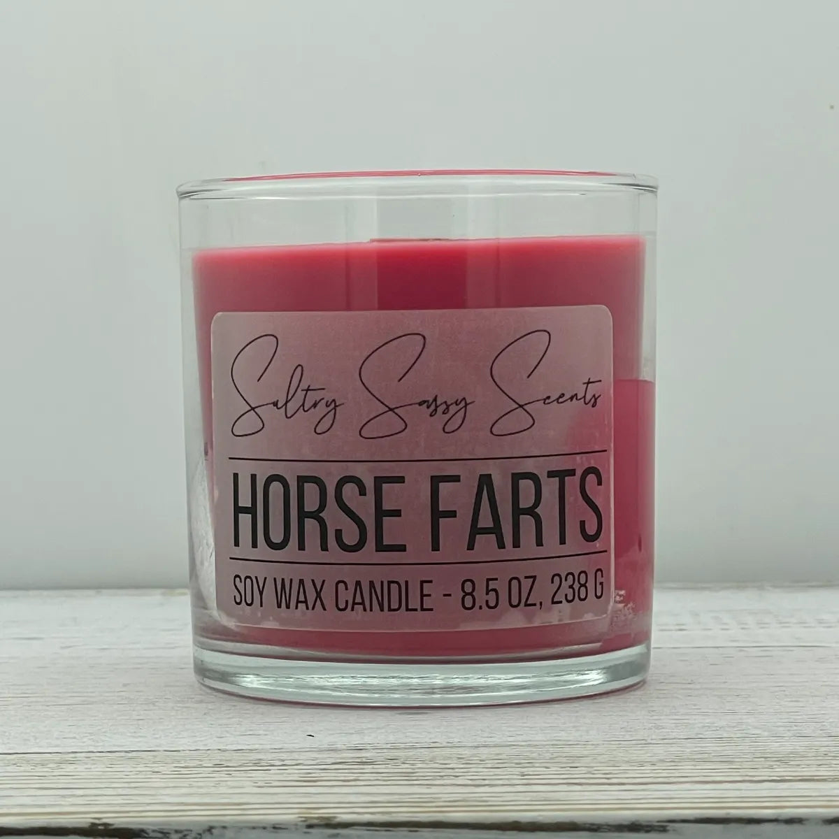 An 8.5-ounce clear glass jar of Sultry Sassy Scents "Horse Farts" soy wax candle showcasing a smooth, deep dark pink wax color with a flat wooden wick.