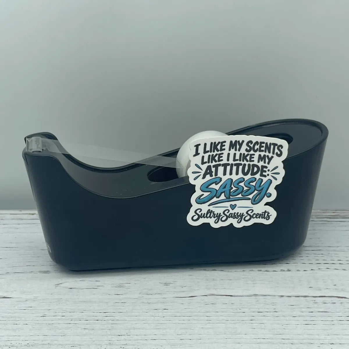 A black desktop tape dispenser holding clear tape, styled with a Sultry Sassy Scents sticker that reads "I like my scents like I like my attitude: Sassy."