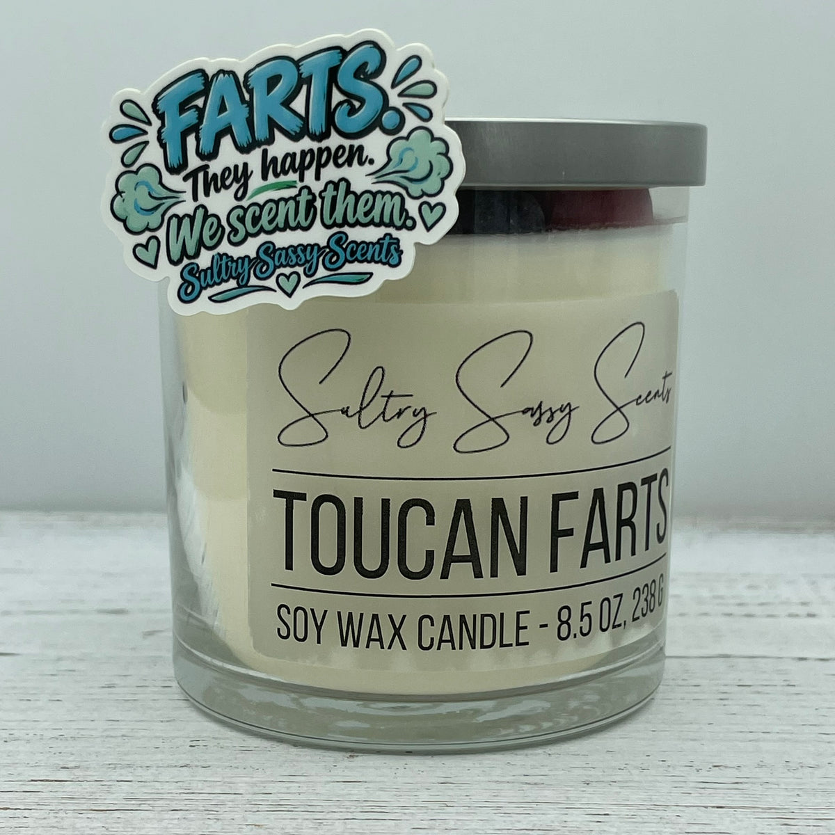 Farts They Happen We Scent Them funny company slogan vinyl sticker showcased next to a Toucan Farts 8.5 oz soy wax candle.