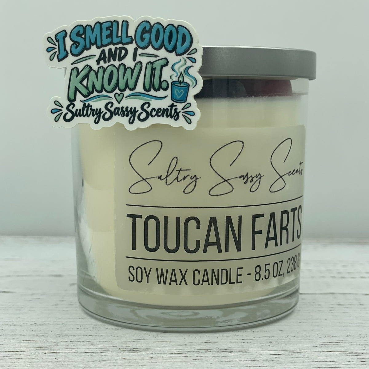 Sultry Sassy Scents I Smell Good and I Know It glossy vinyl sticker propped against a Toucan Farts 8.5 oz wood wick soy candle jar.