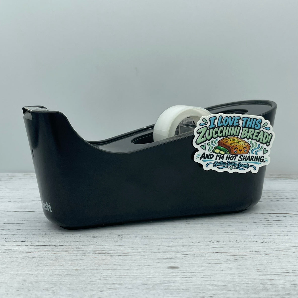 I Love This Zucchini Bread fall bakery vinyl sticker applied to a black plastic tape dispenser on a light rustic wood background.