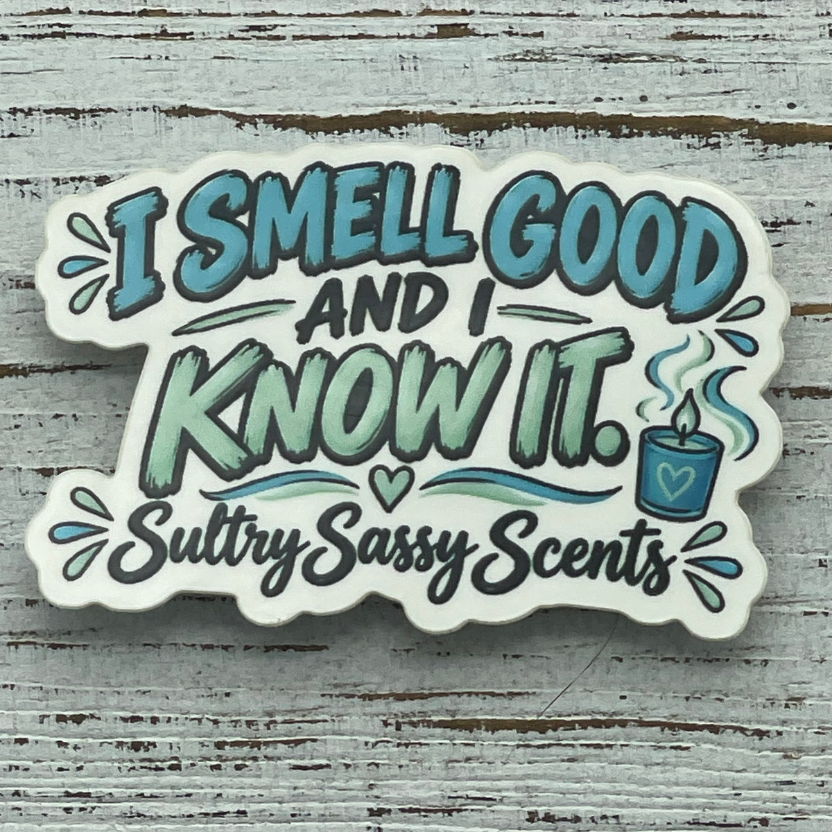 Close-up flat-lay of I Smell Good and I Know It waterproof sticker featuring teal lettering, decorative splashes, and an illustrated burning candle.