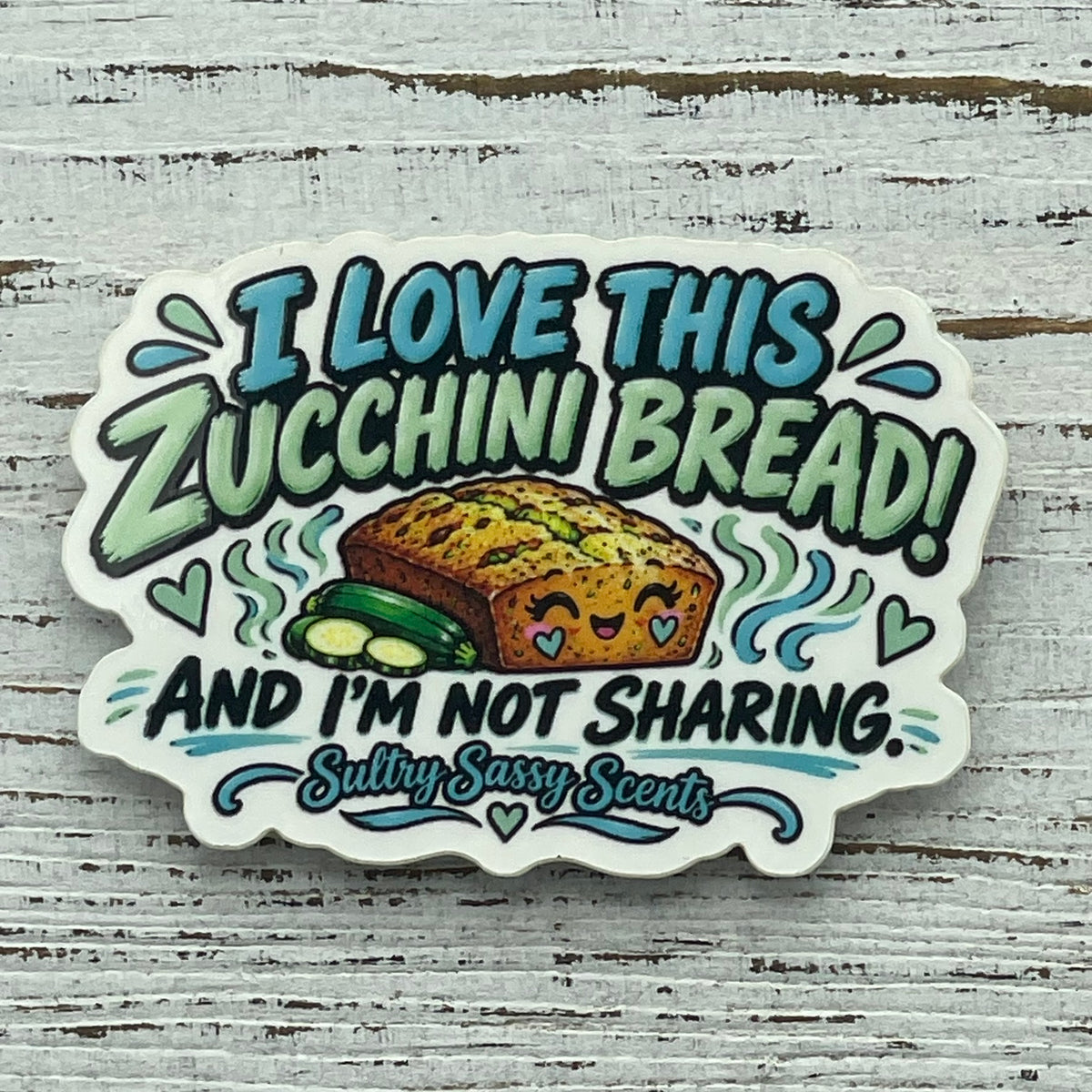 Detailed product shot of a die-cut glossy vinyl sticker featuring a cute smiling zucchini bread loaf illustration with teal and green text layers.