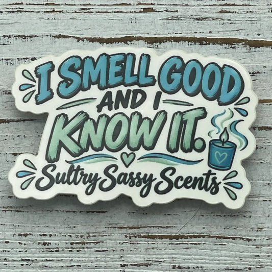 A close-up view of a die-cut vinyl sticker on a white wooden surface, reading "I smell good and I know it. Sultry Sassy Scents" next to a small blue candle graphic.