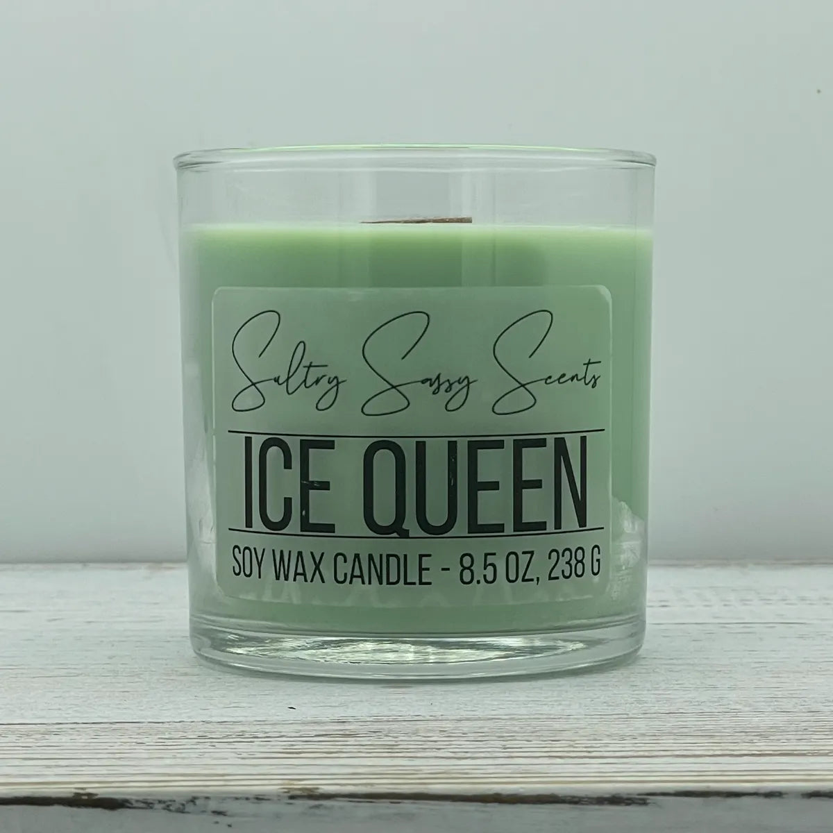 An 8.5-ounce clear glass jar of Sultry Sassy Scents "Ice Queen" soy wax candle featuring a soft pastel mint green wax color and a flat wooden wick, displayed on a rustic light wood table.