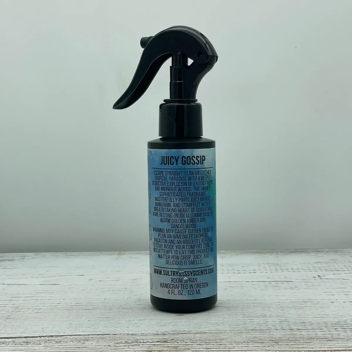Front label of a black spray bottle for Sultry Sassy Scents "Juicy Gossip" Room Spray, showcasing a paragraph-style layout describing a tropical mango and starfruit paradise fragrance.