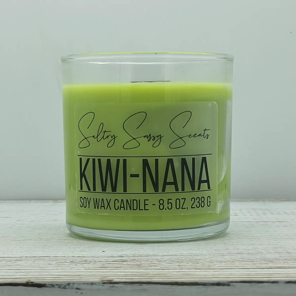 An 8.5-ounce clear glass jar of Sultry Sassy Scents "Kiwi-Nana" soy wax candle displaying a vibrant chartreuse lime green wax color, centered against a light gray background.
