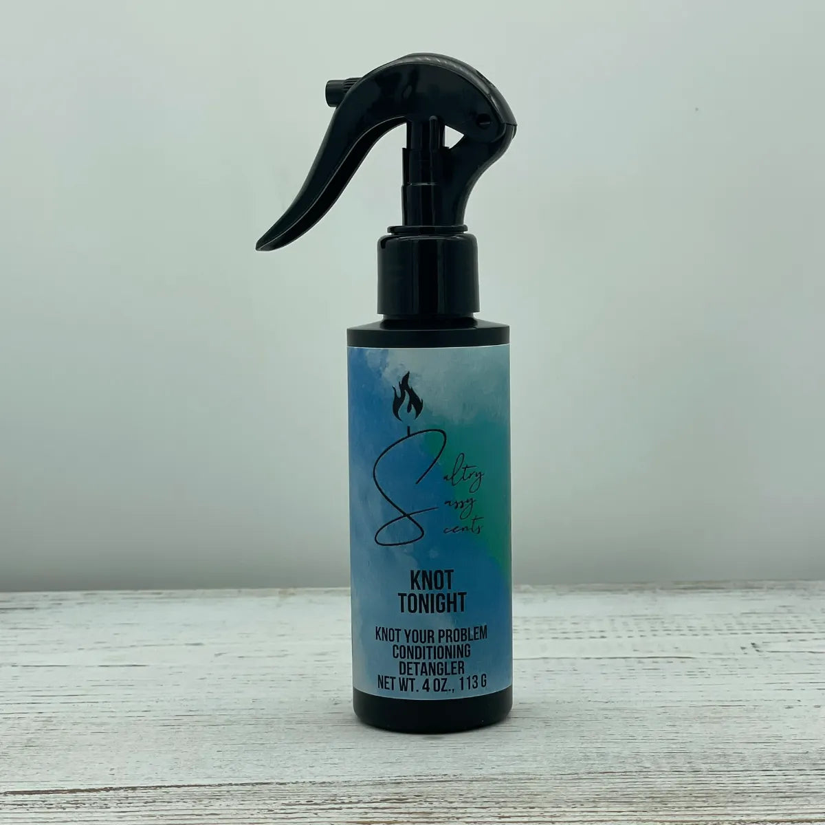 A single black spray bottle of Sultry Sassy Scents Knot Your Problem Conditioning Detangler in the Knot Tonight variation, centered on a light wood background.