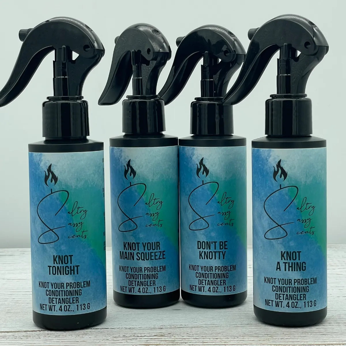 Four black spray bottles of Sultry Sassy Scents Knot Your Problem Conditioning Detangler lined up in a row on a white wooden surface. The variations shown from left to right are Knot Tonight, Knot Your Main Squeeze, Don't Be Knotty, and Knot a Thing.