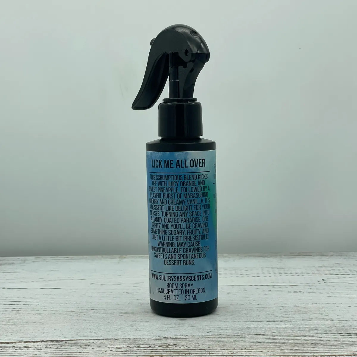 Front label of a black spray bottle for Sultry Sassy Scents "Lick Me All Over" Room Spray, featuring a playful description of a sweet fruity blend with orange, pineapple, and cherry notes.