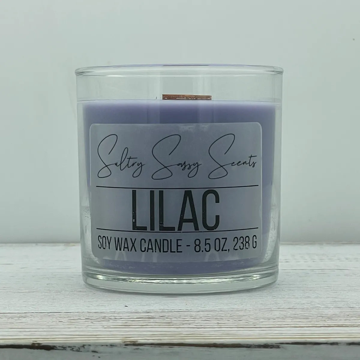 An 8.5-ounce clear glass jar of Sultry Sassy Scents "Lilac" soy wax candle showcasing a smooth pastel lavender purple wax color with a flat wooden wick, resting on a white-washed wood table.