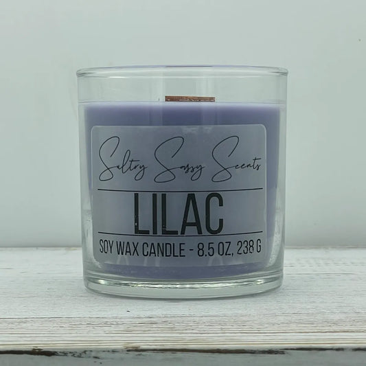 An 8.5-ounce clear glass jar of Sultry Sassy Scents "Lilac" soy wax candle showcasing a smooth pastel lavender purple wax color with a flat wooden wick, resting on a white-washed wood table.