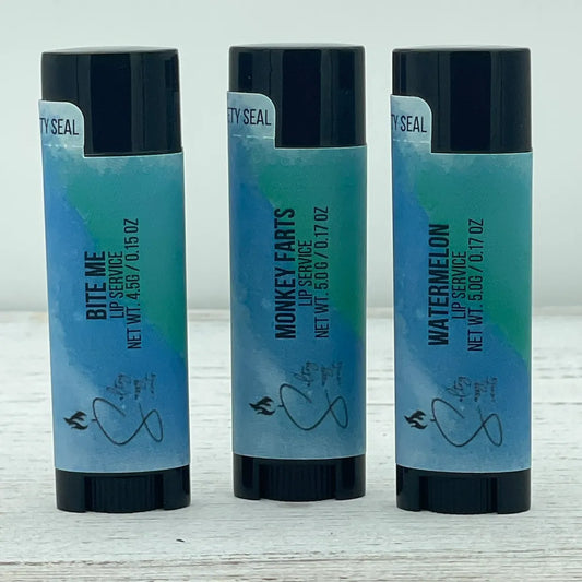  Three black oval tubes of Sultry Sassy Scents Lip Service lip balm lined up in a row on a white wooden surface. The flavor variations shown from left to right are Bite Me, Monkey Farts, and Watermelon.
