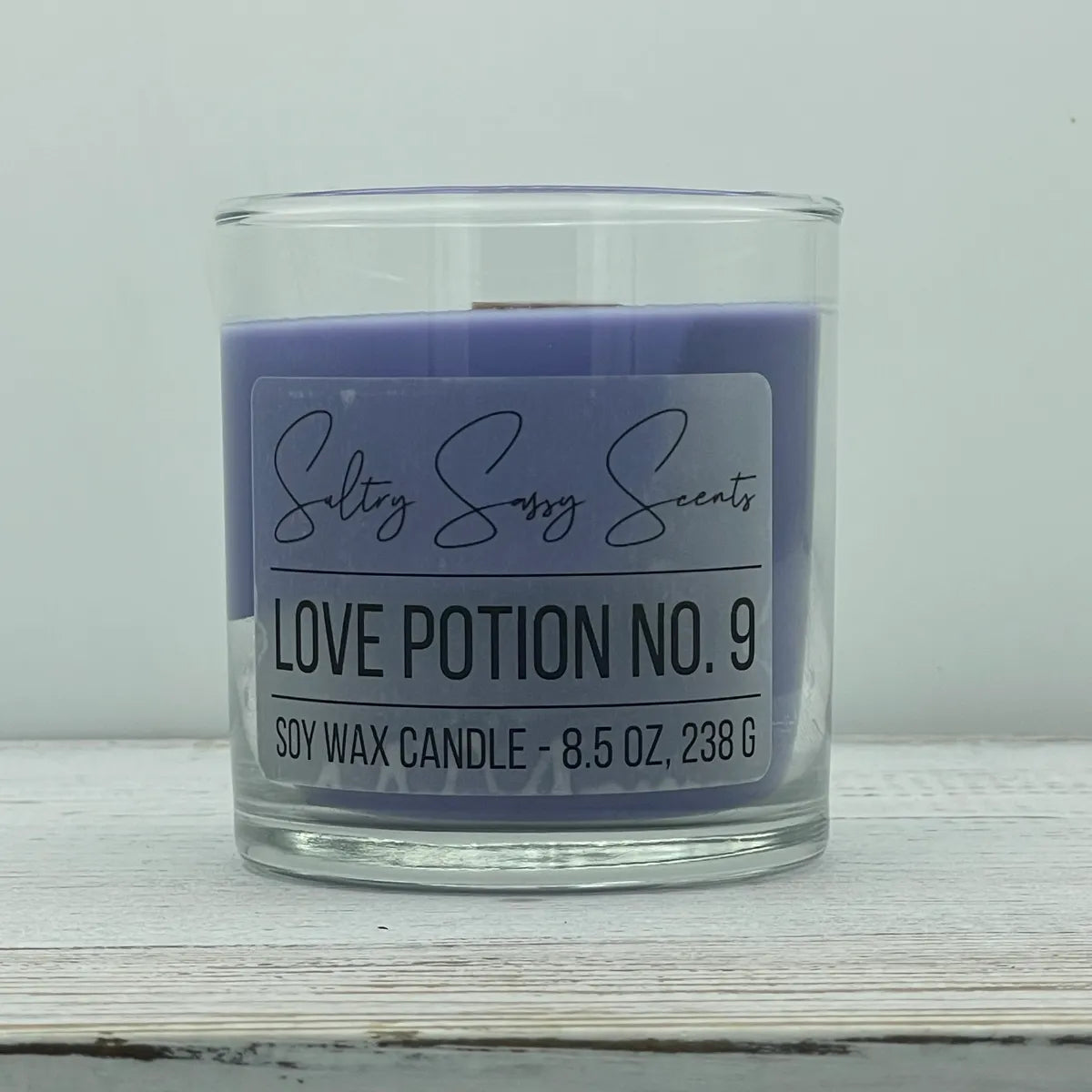 An 8.5-ounce clear glass jar of Sultry Sassy Scents "Love Potion No. 9" soy wax candle featuring a smooth medium purple wax color and a flat wooden wick, displayed on a light wooden background.