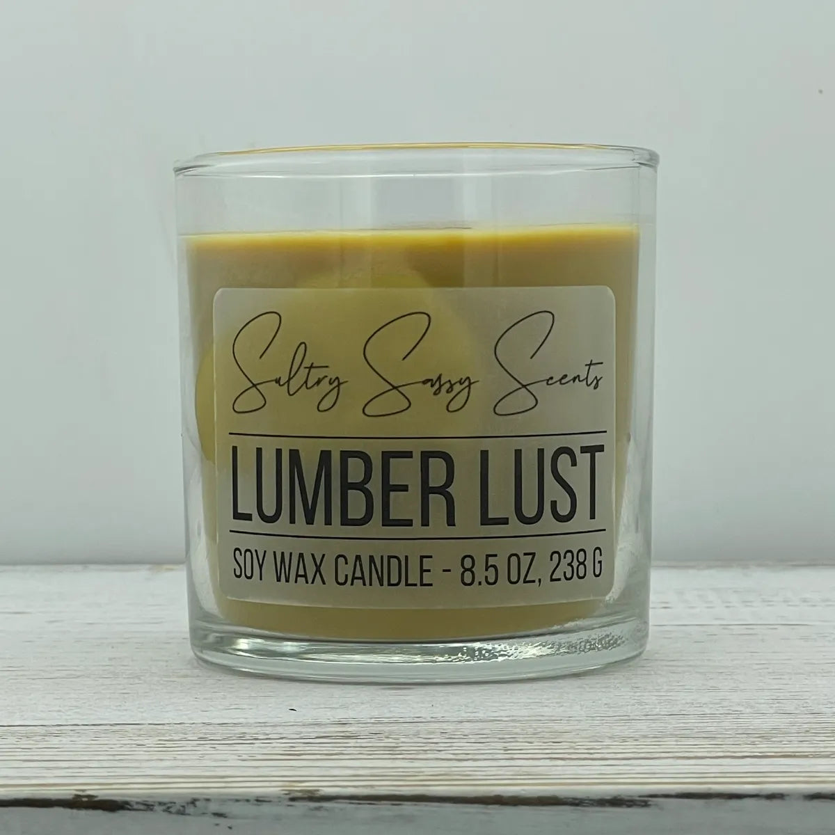 An 8.5-ounce clear glass jar of Sultry Sassy Scents "Lumber Lust" soy wax candle showcasing a deep golden tan wax color with a flat wooden wick, positioned against a clean light background.