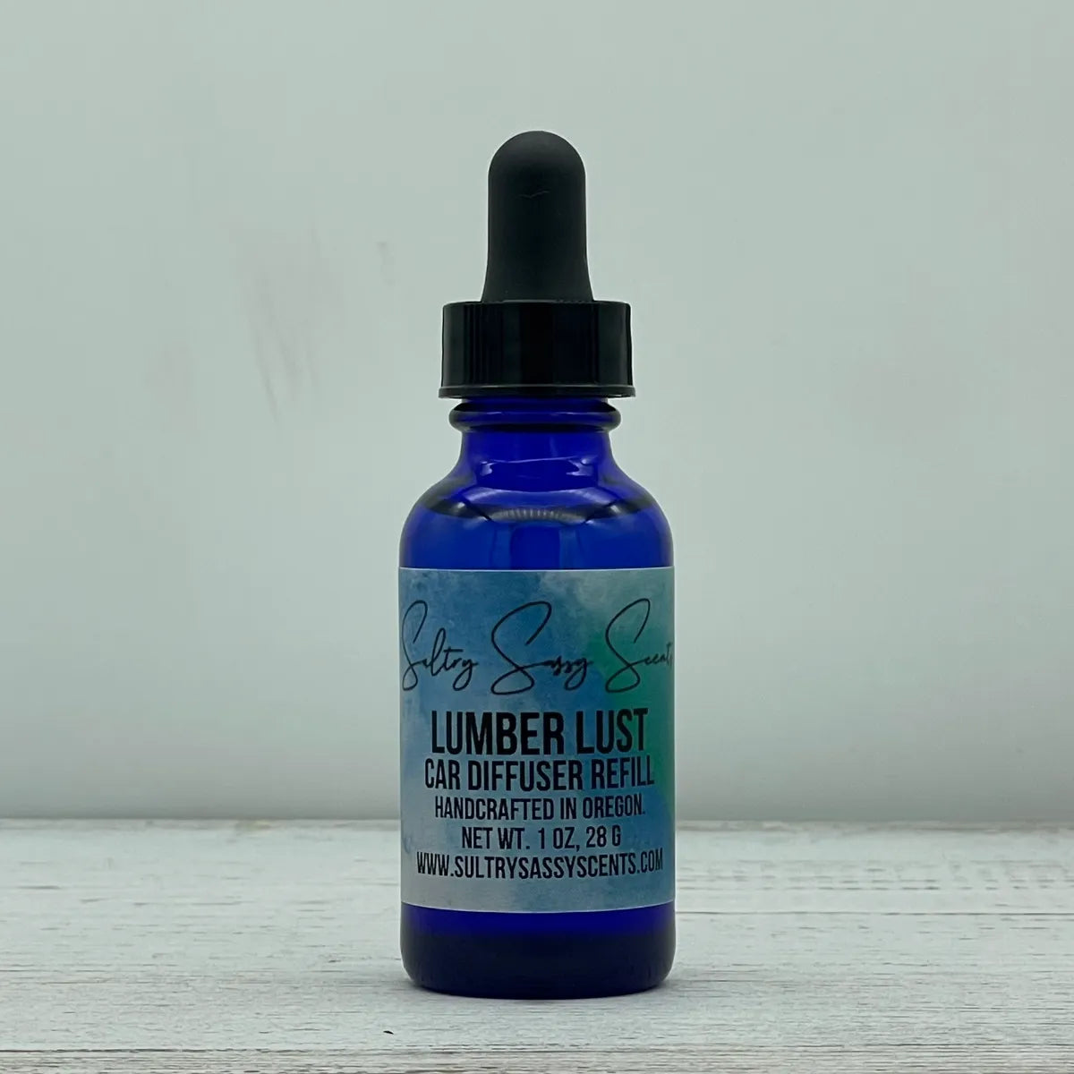 Front label view of a cobalt blue glass dropper bottle containing Sultry Sassy Scents "Lumber Lust" Car Diffuser Refill, detailing the 1 ounce net weight.