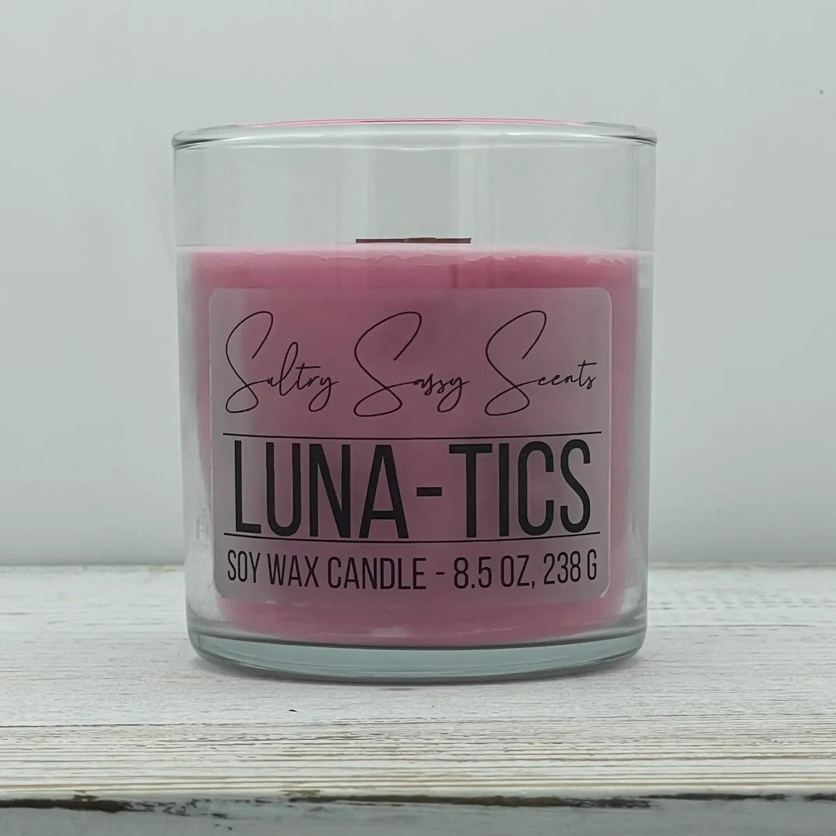 An 8.5-ounce clear glass jar of Sultry Sassy Scents "Luna-Tics" soy wax candle highlighting a soft, dusty rose pink wax color and a flat wooden wick, centered on a light wood surface.