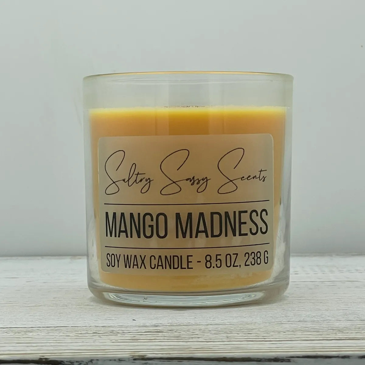 An 8.5-ounce clear glass jar of Sultry Sassy Scents "Mango Madness" soy wax candle featuring a rich mango orange wax color, displayed on a rustic white wood background.