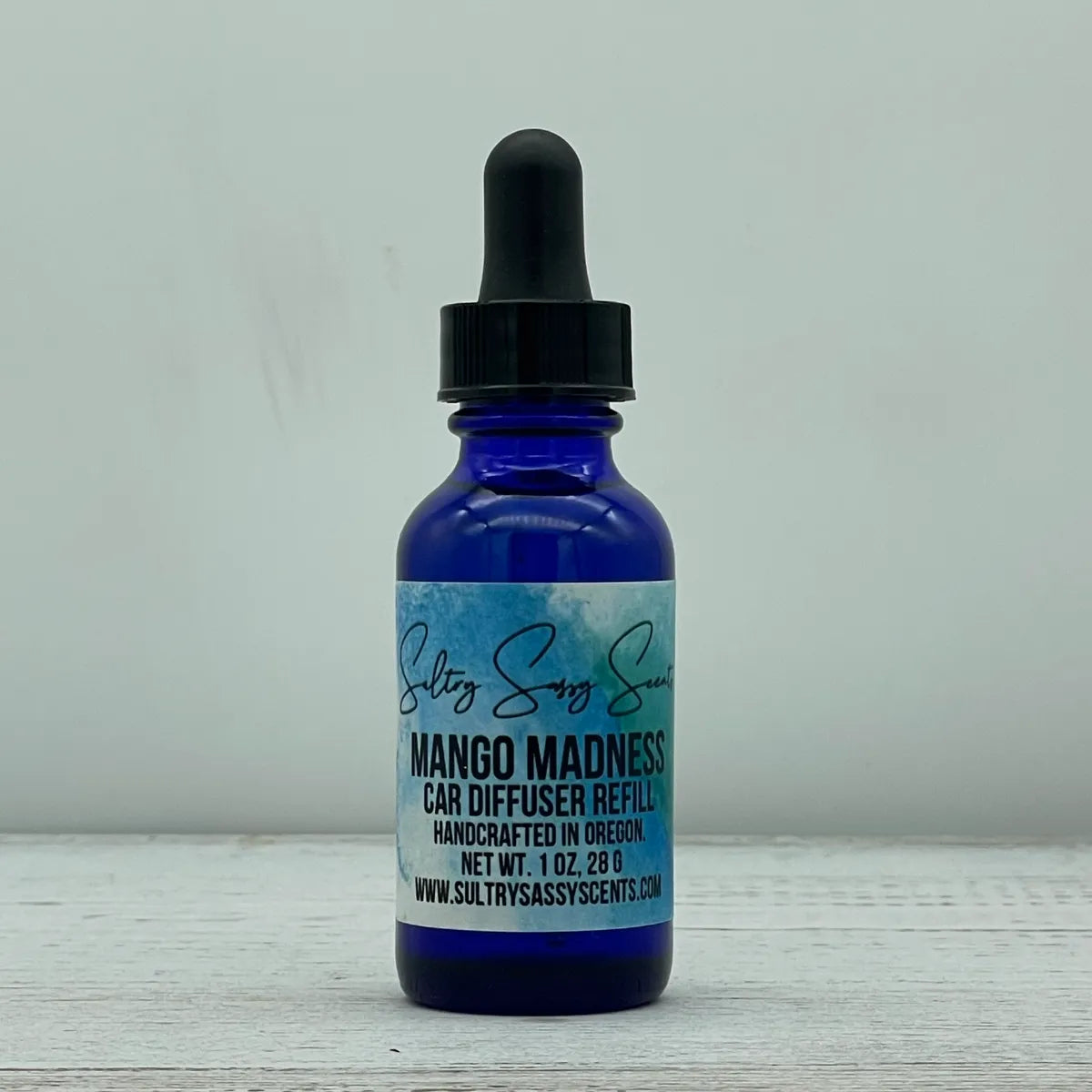 Front label view of a cobalt blue glass dropper bottle containing Sultry Sassy Scents "Mango Madness" Car Diffuser Refill, highlighting the text showing it is handcrafted in Oregon.