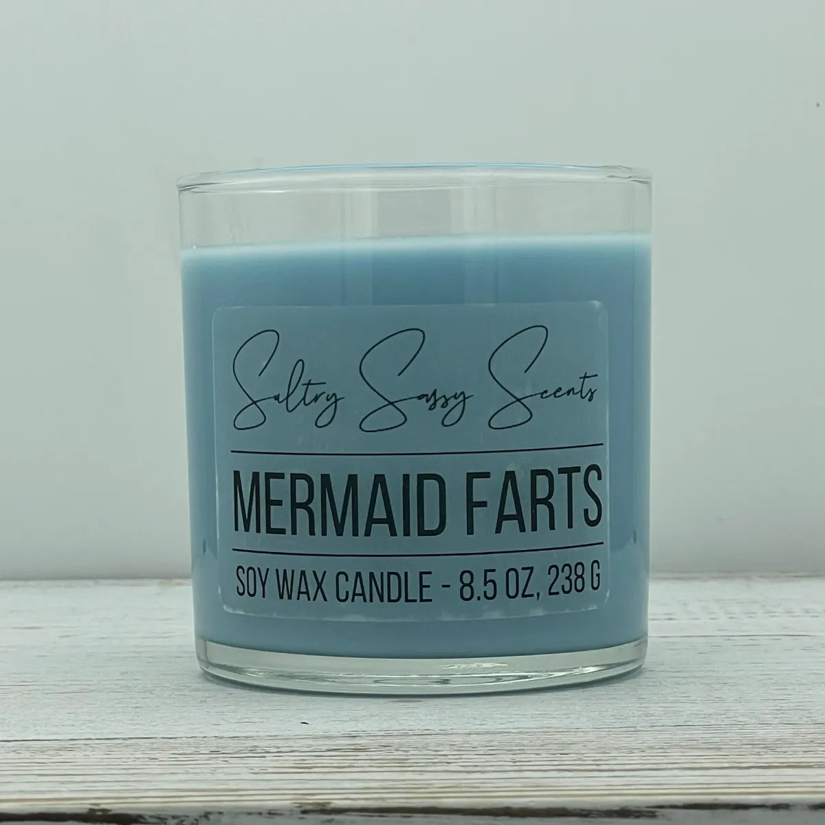 An 8.5-ounce clear glass jar of Sultry Sassy Scents "Mermaid Farts" soy wax candle showcasing a smooth cornflower blue wax color with a flat wooden wick.
