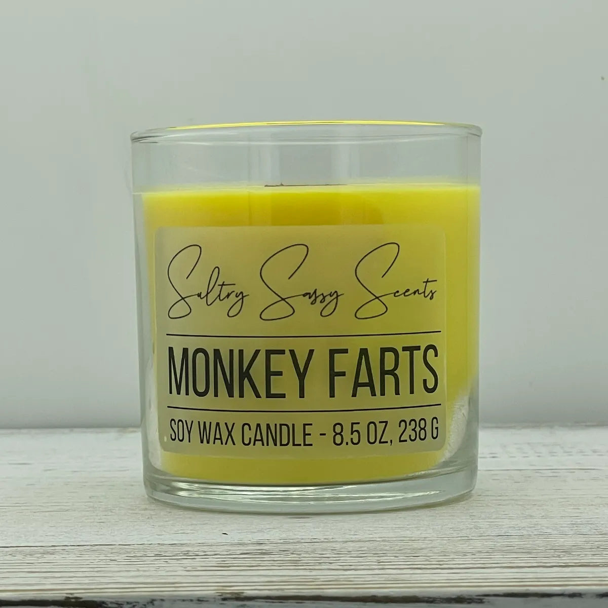 An 8.5-ounce clear glass jar of Sultry Sassy Scents "Monkey Farts" soy wax candle featuring a bright canary yellow wax color and a flat wooden wick, centered on a white wood surface.