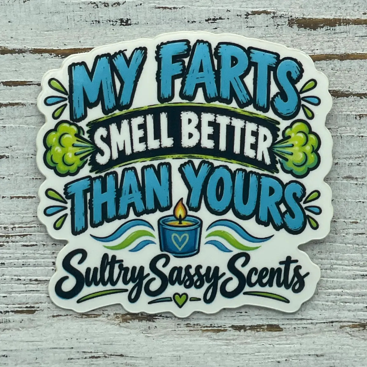 A close-up view of a die-cut vinyl sticker on a white wooden background, reading "My farts smell better than yours. Sultry Sassy Scents" featuring green smoke puff graphics and a small blue candle icon.