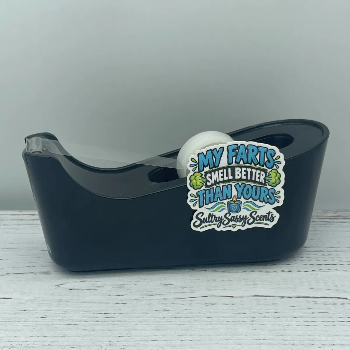 A black plastic tape dispenser sitting on a white wood surface, decorated with a graphic vinyl sticker that says "My farts smell better than yours."