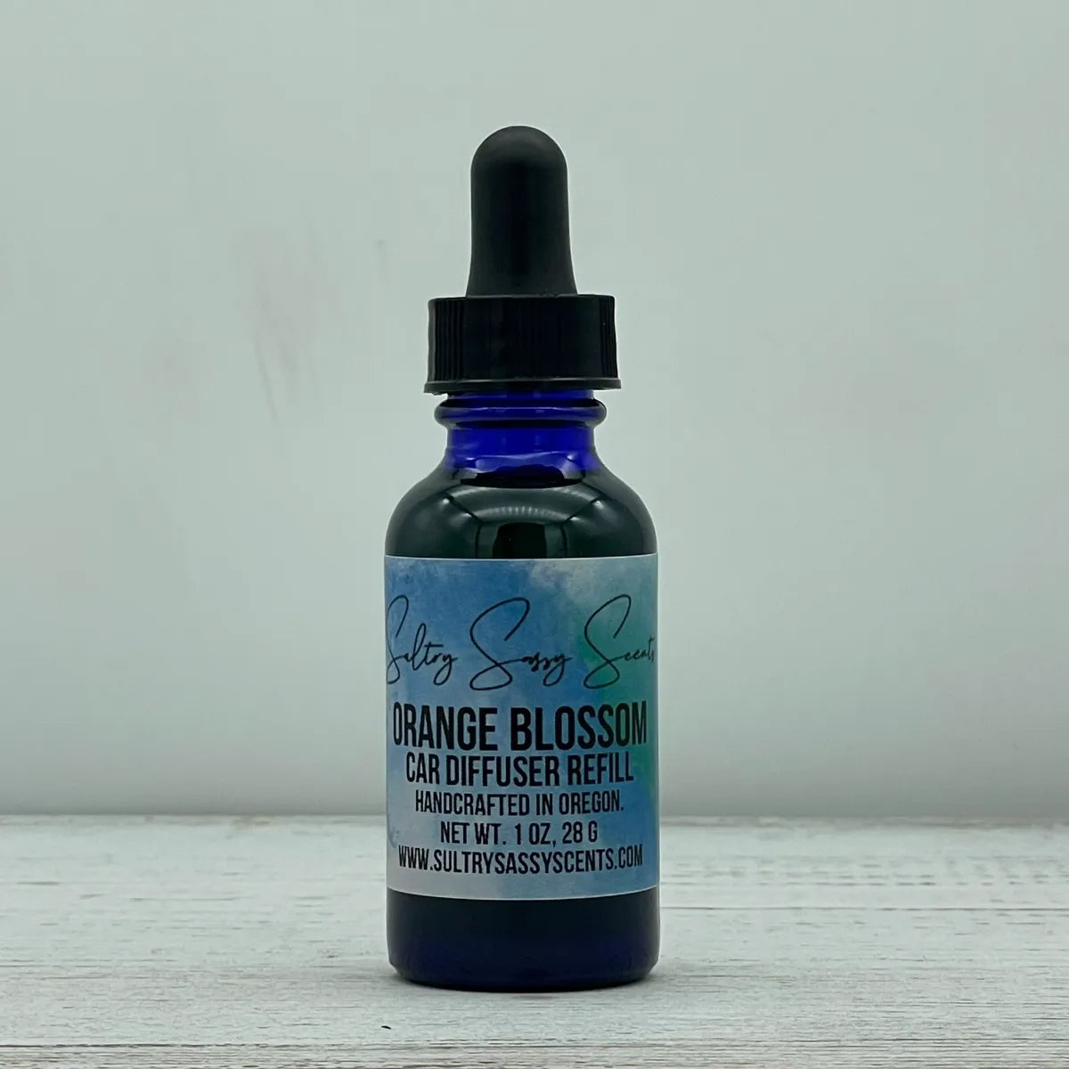 Front label view of a cobalt blue glass dropper bottle containing Sultry Sassy Scents "Orange Blossom" Car Diffuser Refill, centered on a white wood surface.