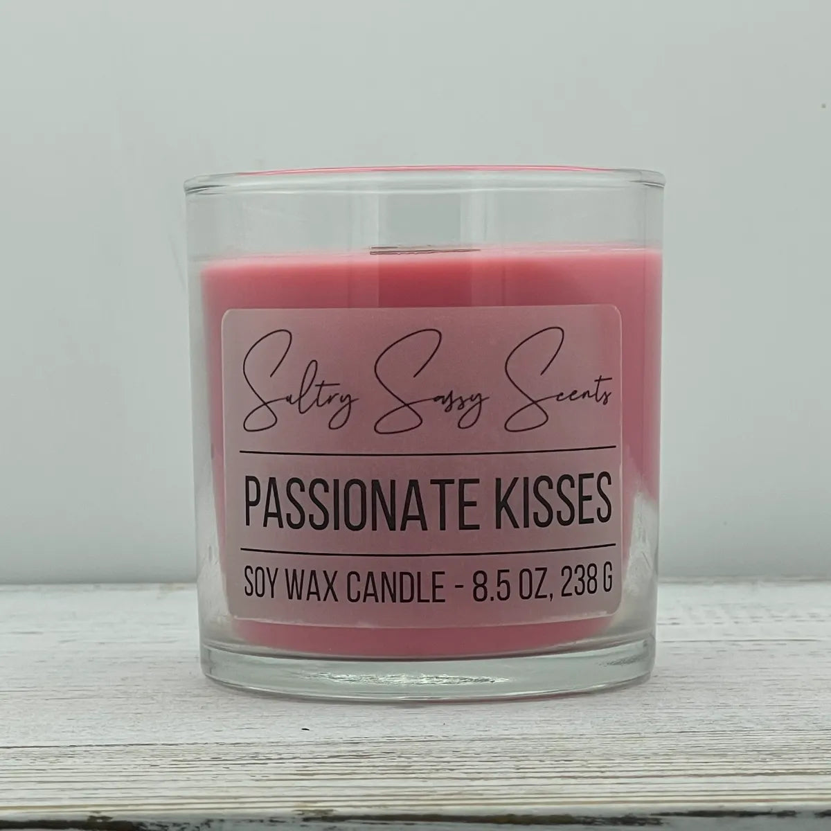 An 8.5-ounce clear glass jar of Sultry Sassy Scents "Passionate Kisses" soy wax candle showcasing a smooth coral pink wax color, centered against a light gray background.