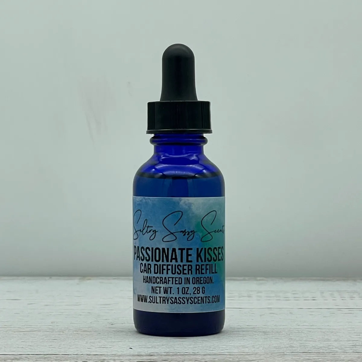 Front label view of a cobalt blue glass dropper bottle containing Sultry Sassy Scents "Passionate Kisses" Car Diffuser Refill, displaying the central brand logo script.