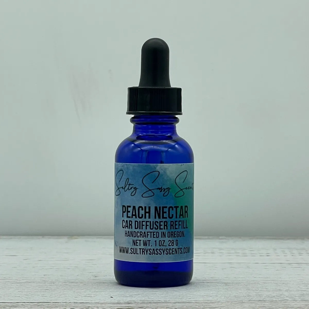 Front label view of a cobalt blue glass dropper bottle containing Sultry Sassy Scents "Peach Nectar" Car Diffuser Refill, positioned on a light wooden background.