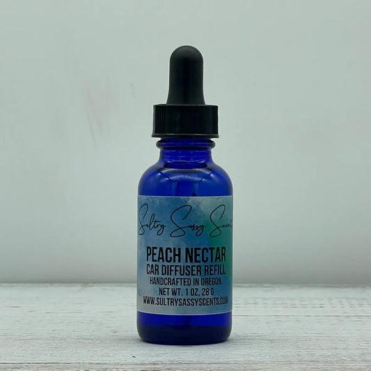 Front label view of a cobalt blue glass dropper bottle containing Sultry Sassy Scents "Peach Nectar" Car Diffuser Refill, positioned on a light wooden background.