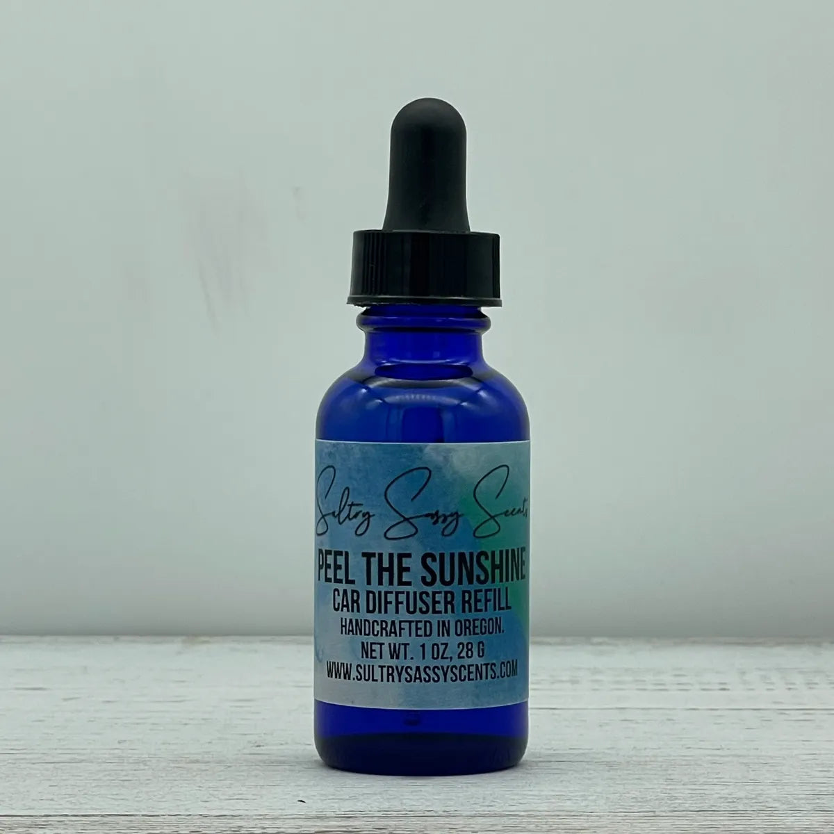 Front label view of a cobalt blue glass dropper bottle containing Sultry Sassy Scents "Peel The Sunshine" Car Diffuser Refill, highlighting the text showing it is handcrafted in Oregon.