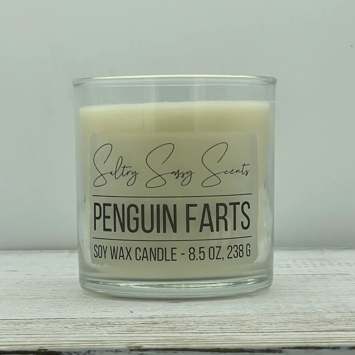 An 8.5-ounce clear glass jar of Sultry Sassy Scents "Penguin Farts" soy wax candle featuring a solid, clean off-white wax color and a flat wooden wick.