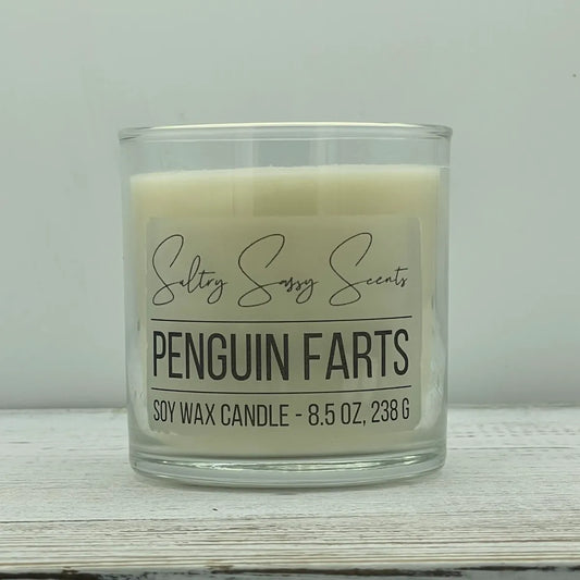 An 8.5-ounce clear glass jar of Sultry Sassy Scents "Penguin Farts" soy wax candle featuring a solid, clean off-white wax color and a flat wooden wick.