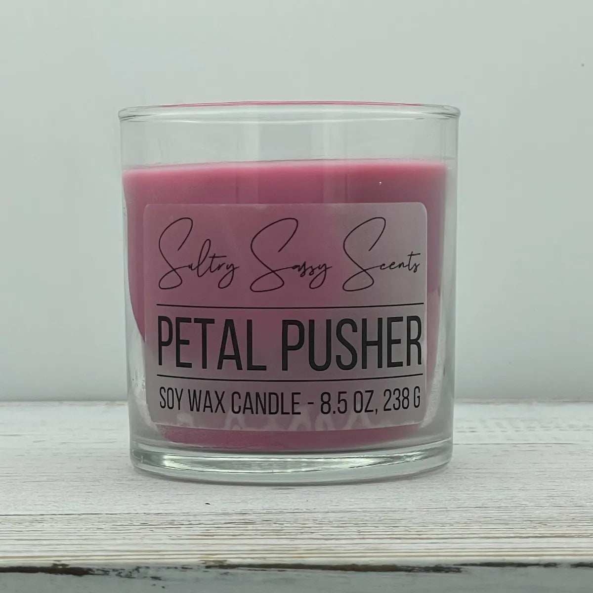 An 8.5-ounce clear glass jar of Sultry Sassy Scents "Petal Pusher" soy wax candle showcasing a vibrant deep pink wax color with a flat wooden wick, resting on a white-washed wood table.