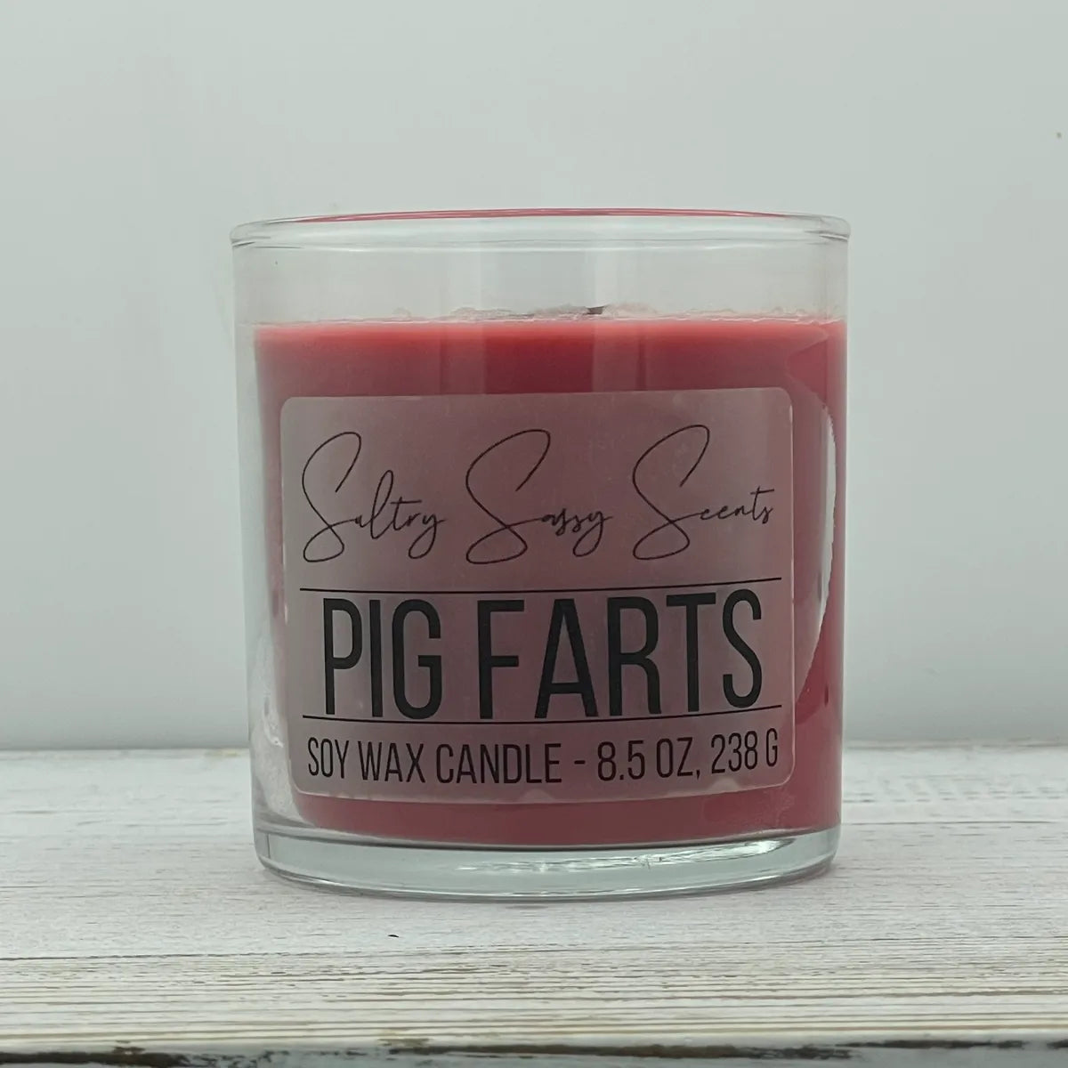 An 8.5-ounce clear glass jar of Sultry Sassy Scents "Pig Farts" soy wax candle displaying a muted plum red wax color, displayed on a light wooden background.