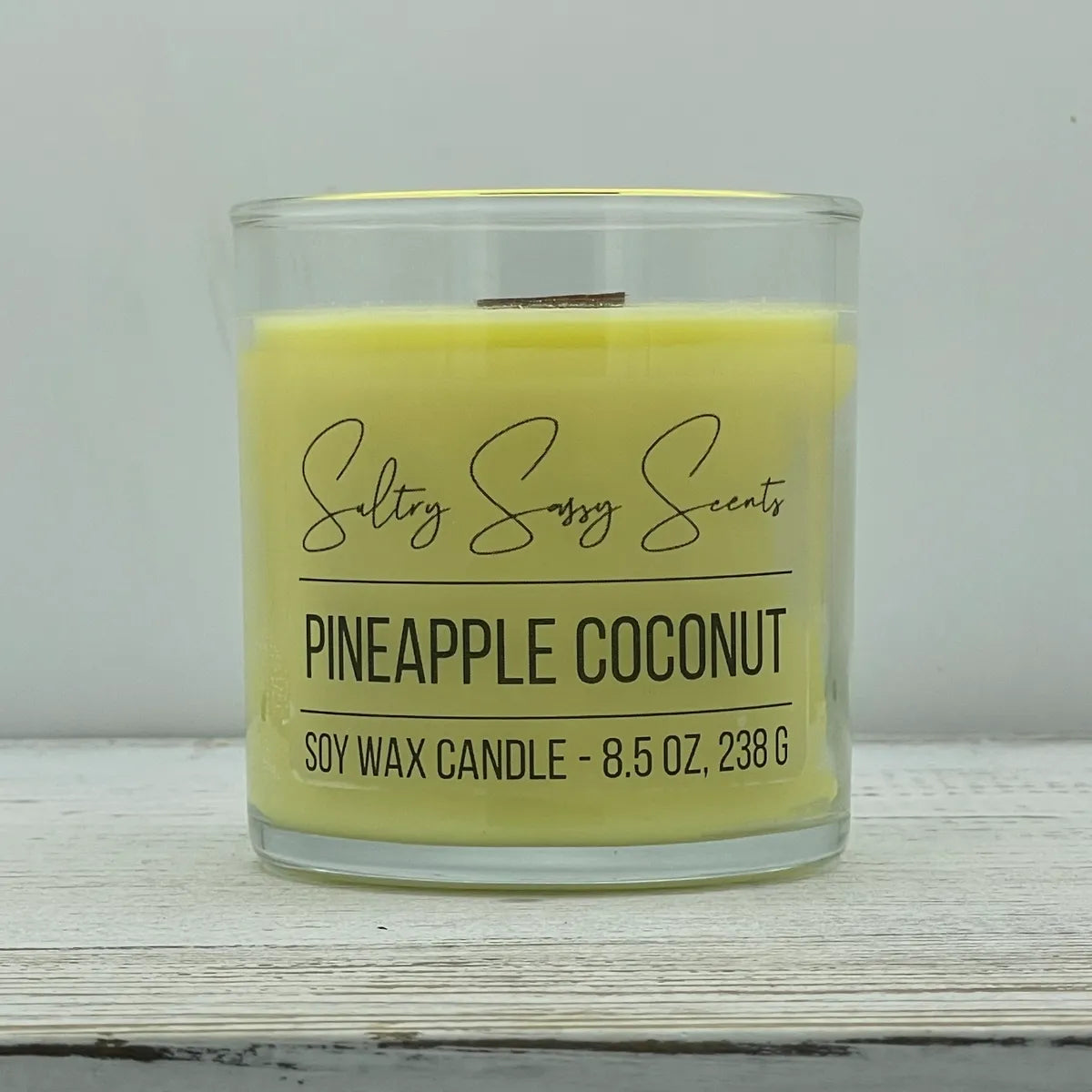 An 8.5-ounce clear glass jar of Sultry Sassy Scents "Pineapple Coconut" soy wax candle featuring a soft pastel yellow wax color and a flat wooden wick.