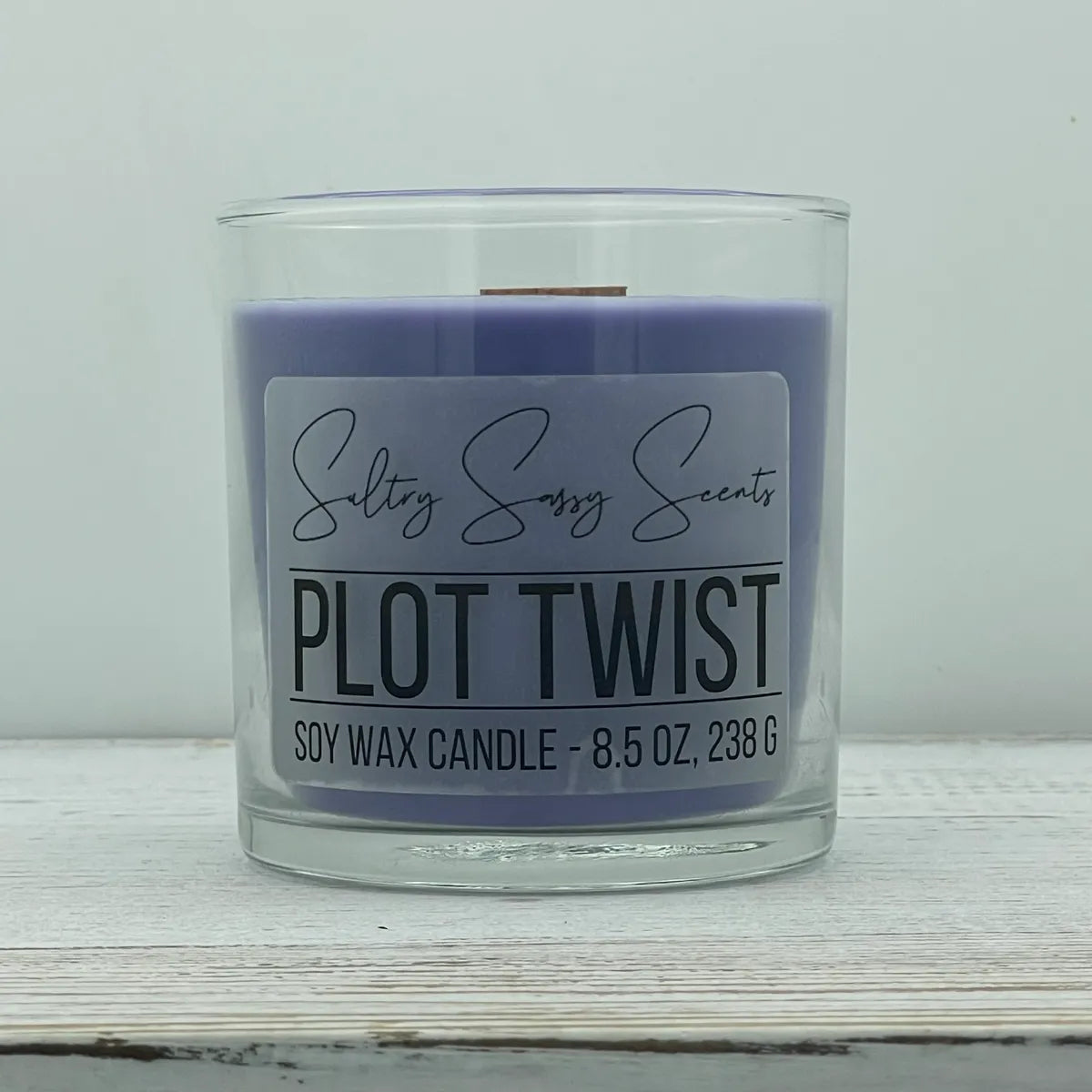 An 8.5-ounce clear glass jar of Sultry Sassy Scents "Plot Twist" soy wax candle showcasing a smooth pastel lavender purple wax color with a flat wooden wick, positioned against a clean light background.