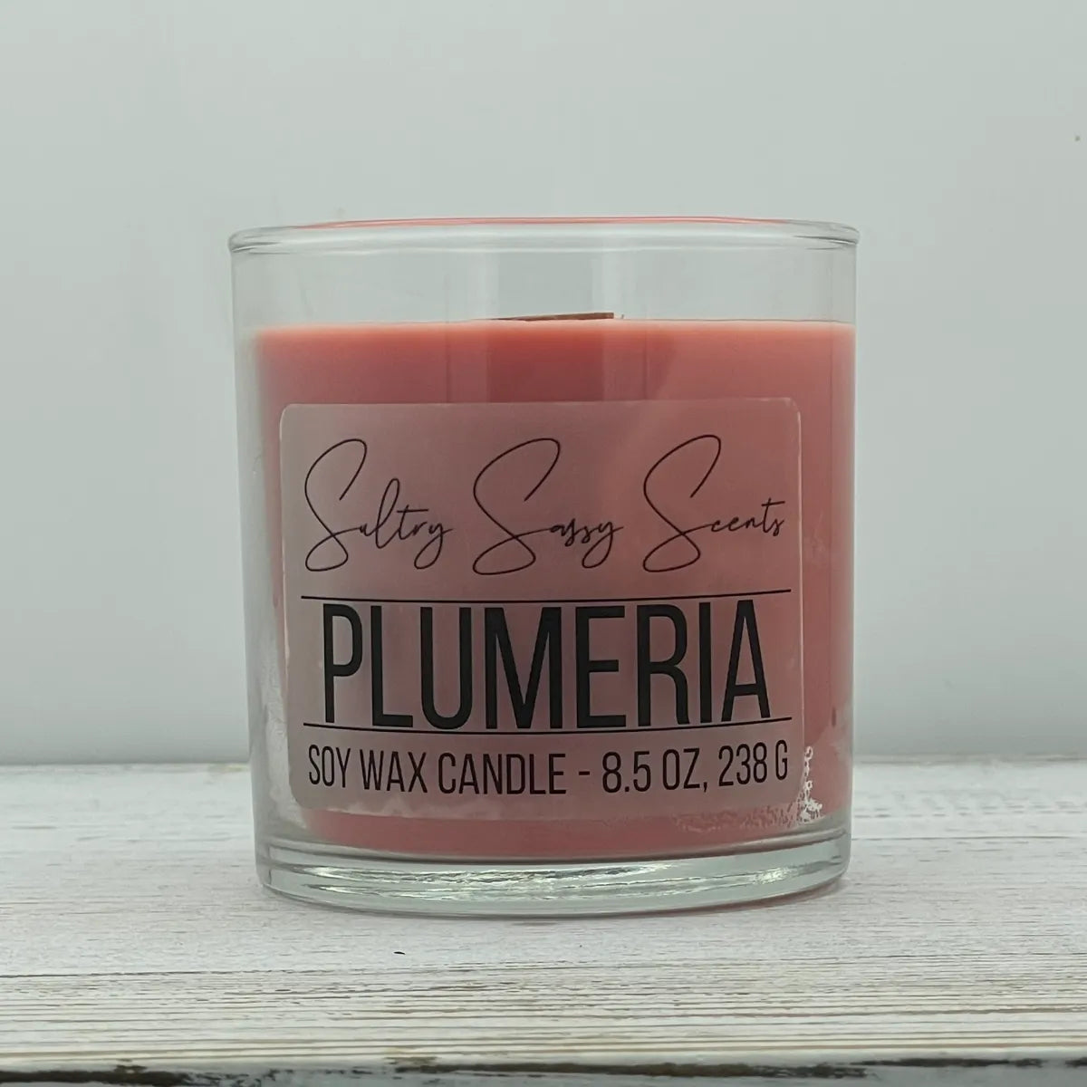 An 8.5-ounce clear glass jar of Sultry Sassy Scents "Plumeria" soy wax candle featuring a soft coral pink wax color, positioned against a clean light backdrop.